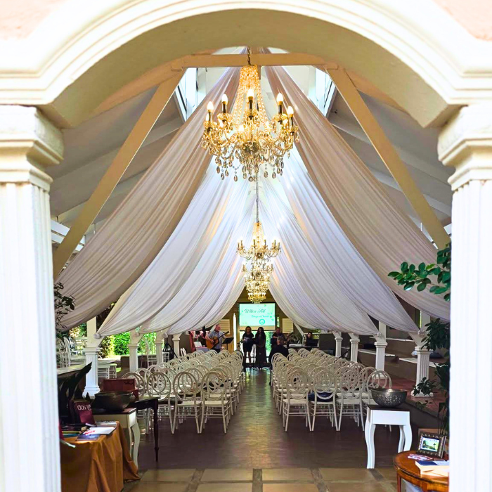 A wide archway view into an elegant hall decorated with flowing white drapery, chandeliers, and rows of tables set for an event.