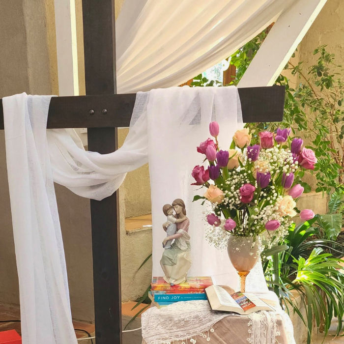 A wooden cross draped in white fabric with flowers, a small figurine, and an open Bible arranged at its base.