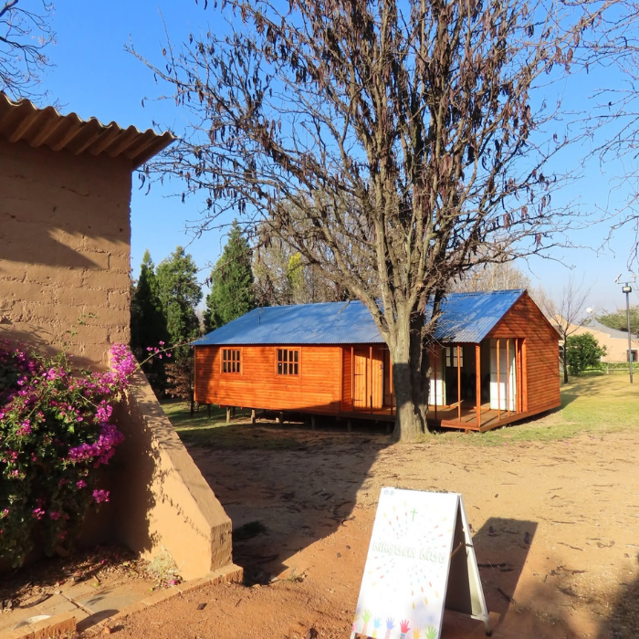 Two wooden cabins with blue roofs standing under a bare tree on a dirt property, with bougainvillea flowers in the foreground.