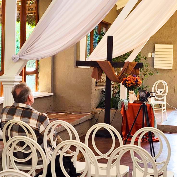 A man seated among white chairs facing a wooden cross draped in white fabric with flowers, in an indoor worship setting.
