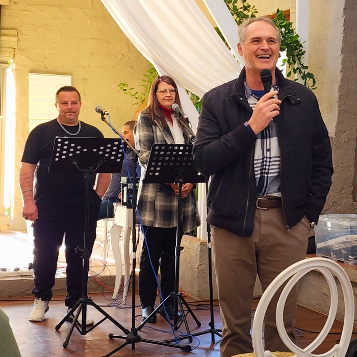 Three worship team members singing and speaking into microphones on stage during a church service, with a draped cross behind them.
