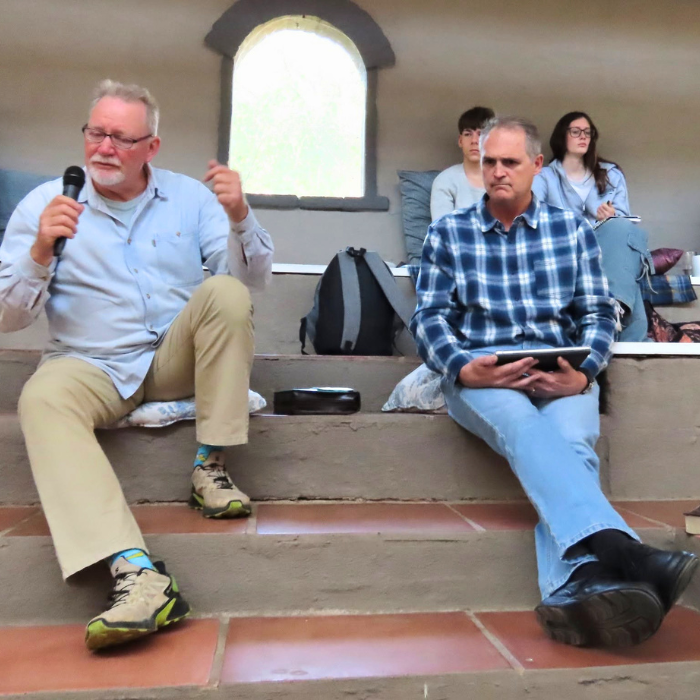 A man holding a microphone speaking while seated on stone steps, with others listening nearby.