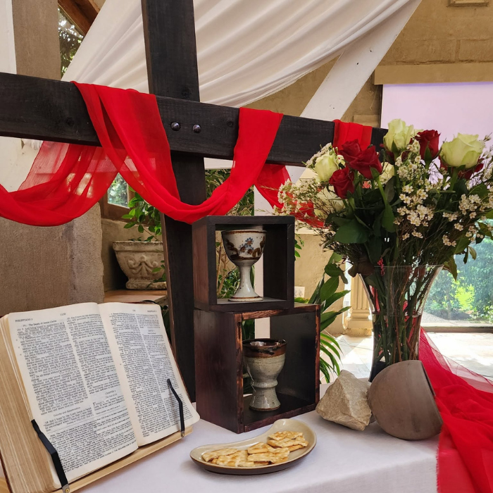 A communion table with an open Bible, wafers, a chalice, wine, and red and white flowers, set beneath a wooden cross draped in red fabric.
