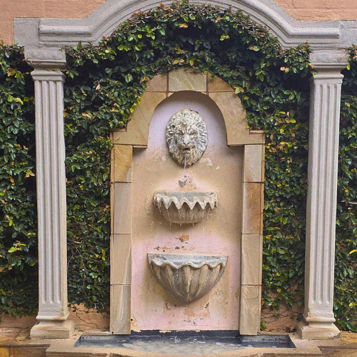 A decorative stone fountain with a lion's head, framed by stone columns and covered in green ivy.