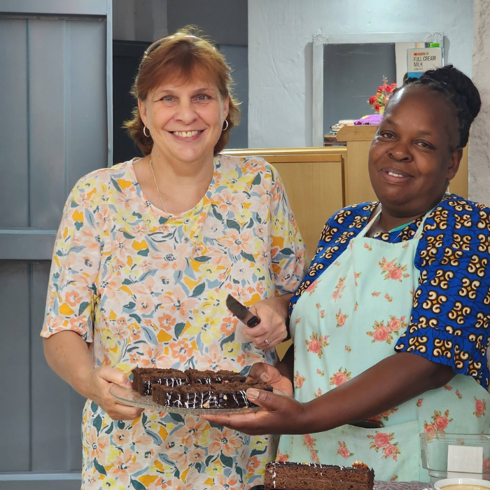 Two smiling women holding a freshly baked chocolate cake together in a kitchen.