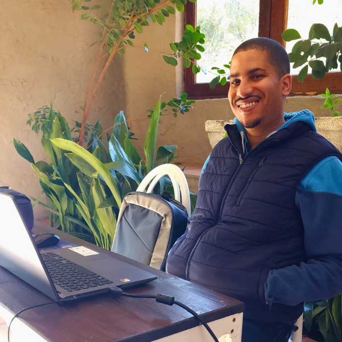 A smiling man seated beside a laptop and audio equipment on a wooden table, with plants in the background.