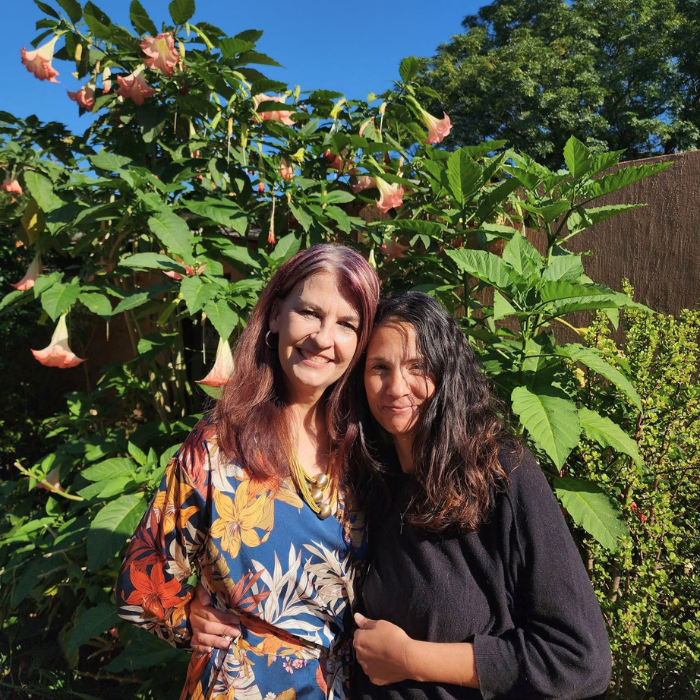 Two smiling women standing together in front of tall pink flowering plants and greenery.