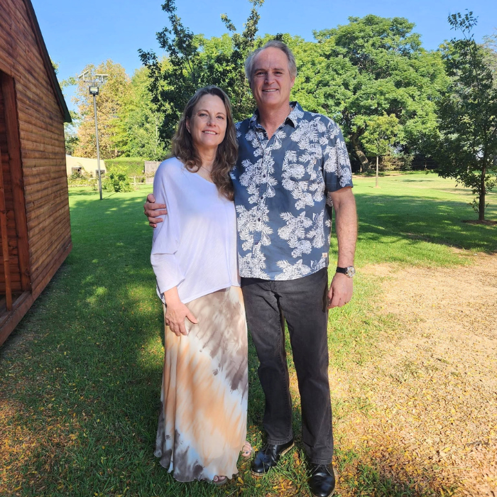 A smiling couple standing together on a green lawn, surrounded by trees.