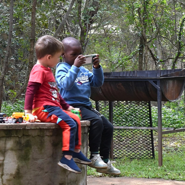 Two young boys sitting on a stone wall outdoors, one filming with a phone, near a fire pit and trees.
