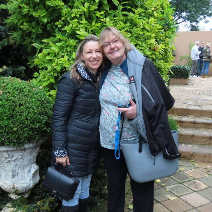 Two women smiling and embracing outdoors in a lush garden, with stone urns and greenery around them.
