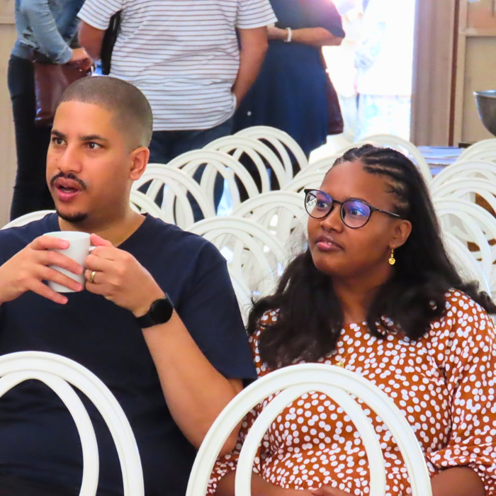 A man holding a mug and a woman seated together, chatting during a gathering with white chairs around them.
